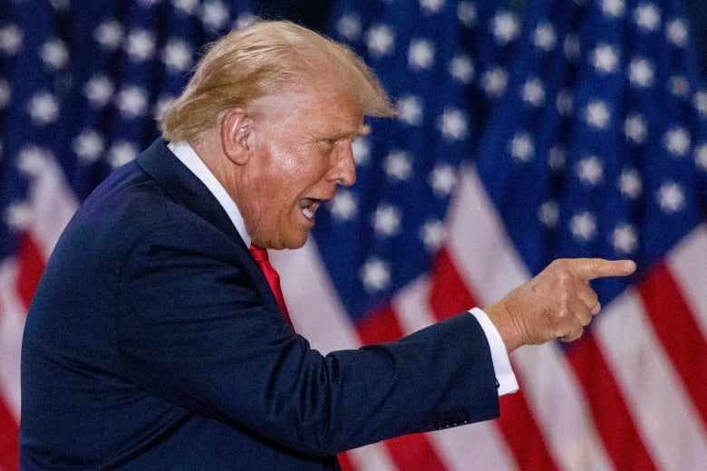 Donald Trump, pictured in profile in front of an American flag, points his finger and speaks.