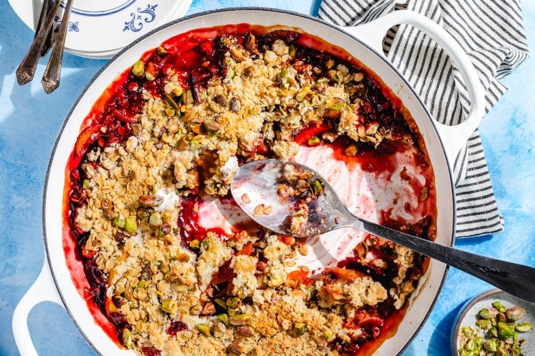 Overhead shot of a white dish of stone fruit crumble topped with pistachios. The crumble has a few spoonfuls missing and a silver spoon sitting in the middle.