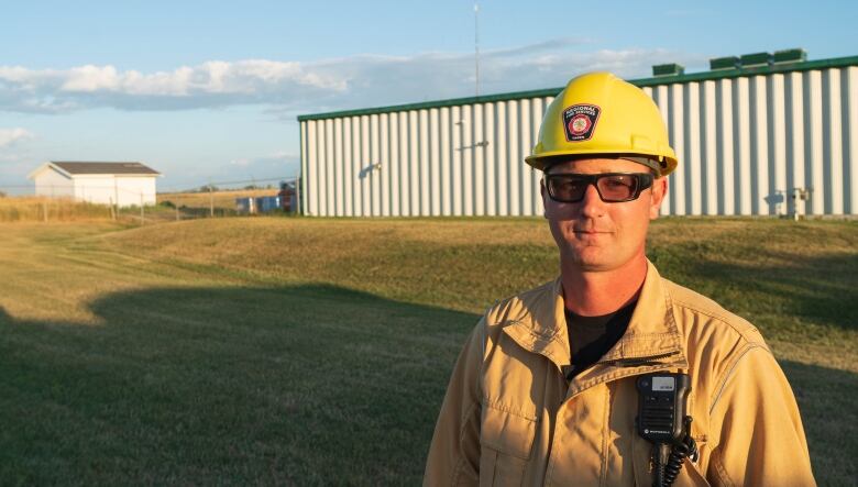 A man wearing a yellow helmet and coveralls stands in front of a warehouse on the grass in the fading afternoon light. 