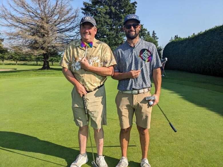 Two men in button-down t-shirts, cackey shorts, white sneakers and ballcaps are holding golf clubs in one hand and Pride accoutrements in the other.