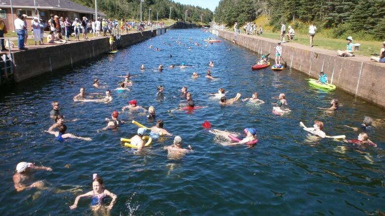 The photo shows a group of swimmers in the canal, surrounded by wooded areas.