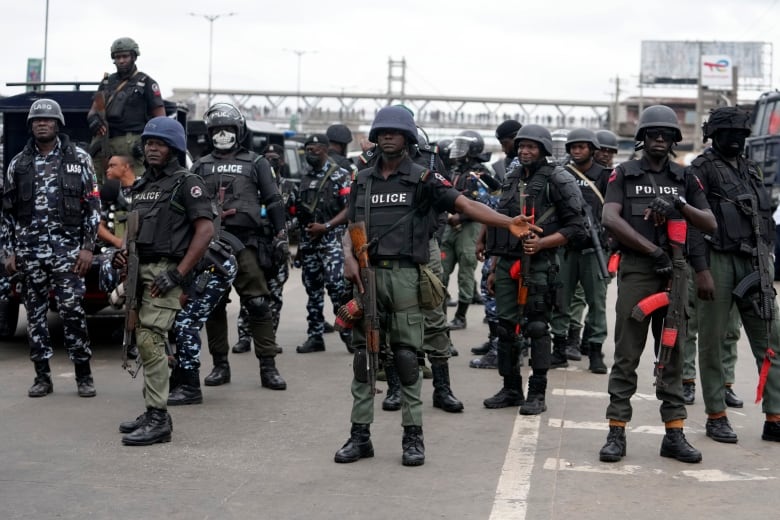 Police with helmets and assault rifles stand on a paved street.