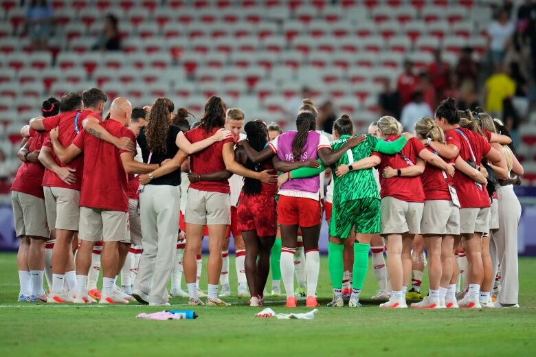 A group of soccer players and coaches stand in a circle arm in arm. 