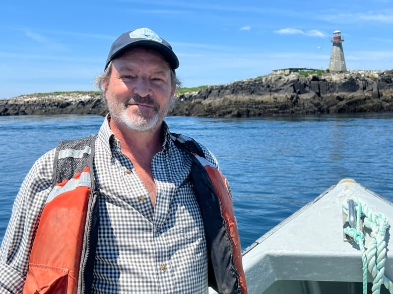 A man with a grey scruffy beard smiles at the camera. he wears a life jacket standing on a boat. In the background is a lighthouse.