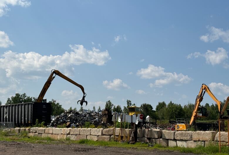 Cranes moving material with train cars to the left and a wall of large cement blocks in the foreground.