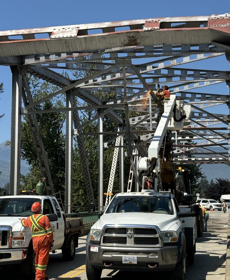 Crews repair a bridge on a sunny day, with one of them using a truck lift to access the cross beams.