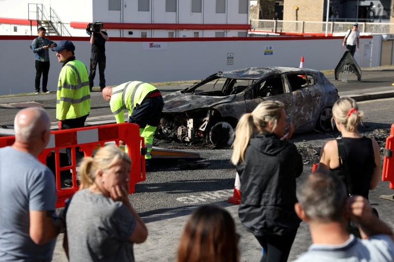 Onlookers gather near a burnt and destroyed car.