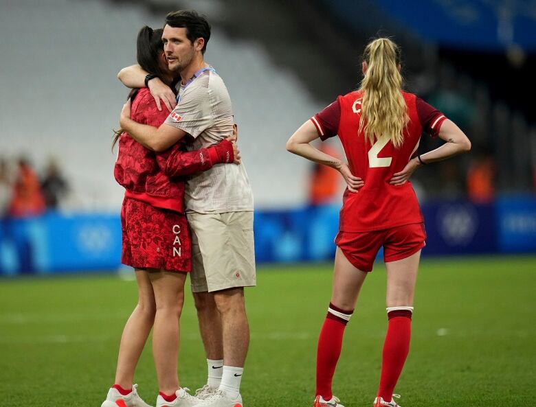 A man wearing Team Canada clothes hugs a female soccer player on a soccer pitch, while another player looks onward.