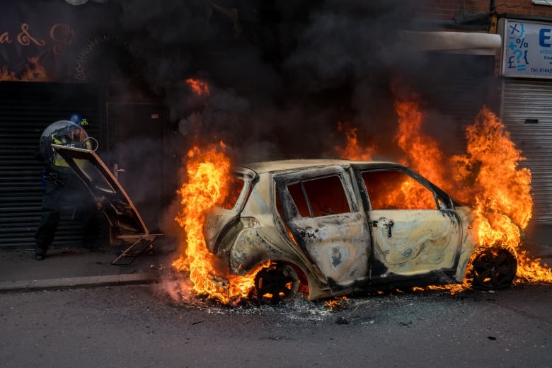 A police officer is riot gear reacts to a burning car.