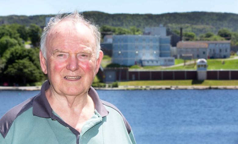 A smiling older man stands in front of lake. On the other side of the lake is a prison.