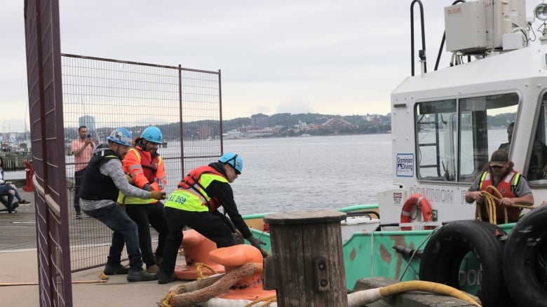 Three people wearing helmets pull on a line from a boat to the right. Another person is on the boat holding the line.