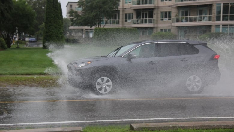 A grey car drives through water pooled on a roadway, splashing water up into the air