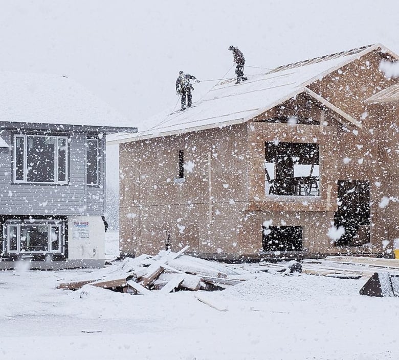 Two people work on the roof of a wood house structure under construction with snow blowing around them. 