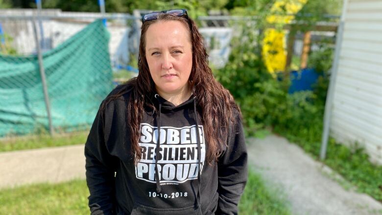 A woman stands in a yard. She is wearing a black hoodie with the words 