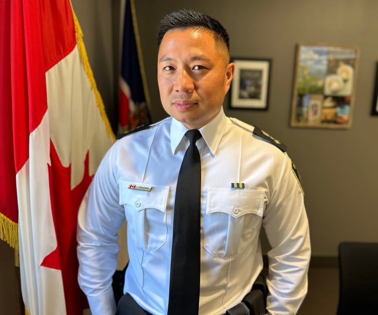 London's police chief in a white dress shirt stands and stares at the camera with a Canadian flag in the background. 