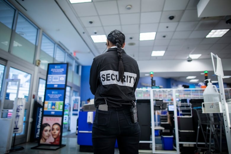 A female security guard with a long braid in her hair and the word Security on the back of her shirt is seen from behind. 