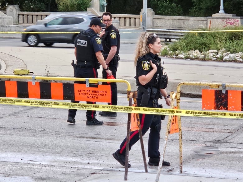 Police officers in black uniforms walk among yellow tape and barricades on a street.