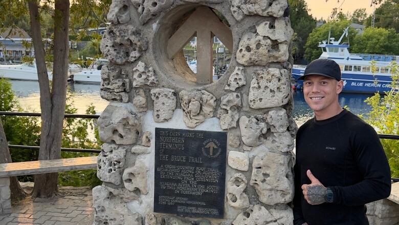 A person in shorts and a hat poses by a stone monument with a sign saying it marks the Bruce Trail.