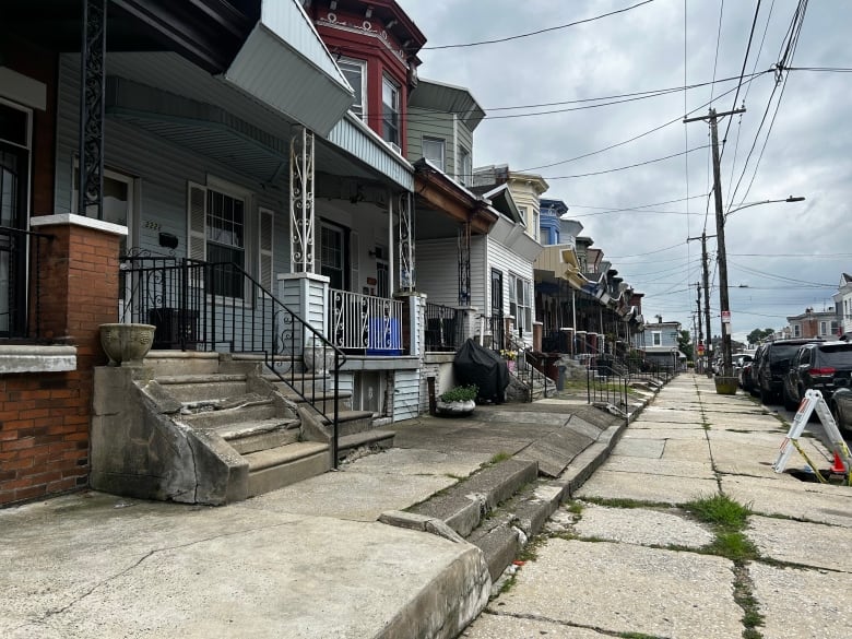 The photos shows a street full of row homes, which are homes that are side by side and share a common wall. The neighbourhood is rundown. There is grass growing out of the cracks in the sidewalk.