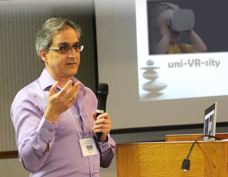 A man holds a microphone and speaks to a crowd out of frame. A projecter behind him displays a picture of a child with a virtual reality headset and the phrase 