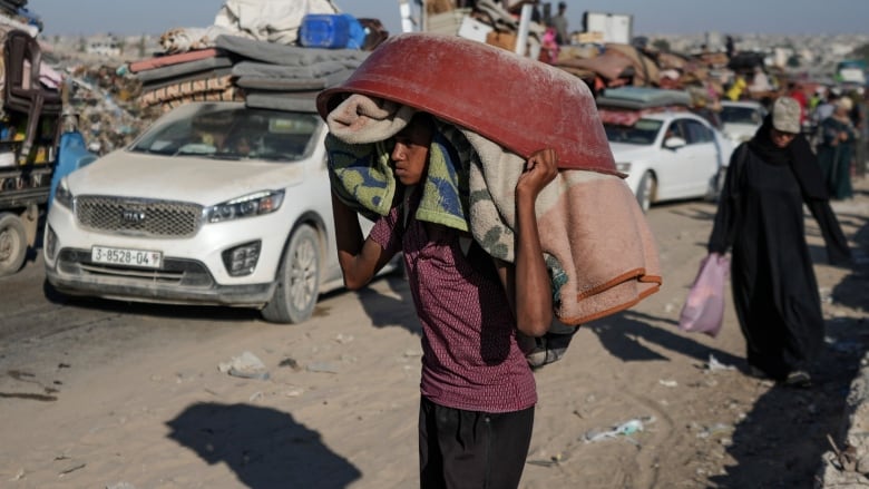 A youth holds a basin and what appear to be a blanket and a towel over their head as they walk on a dusty street.