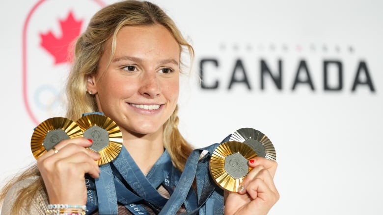 A woman smiles while holding up three Olympic gold medals and one Olympic silver medal that hang around her neck.