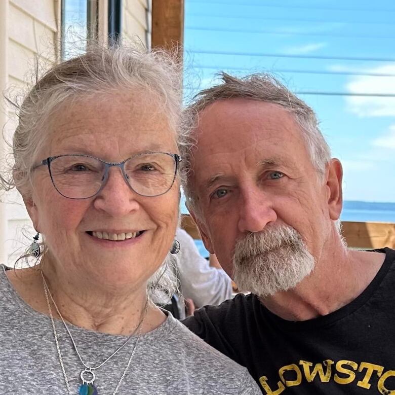 A woman with grey hair and glasses is shown in a photo with her husband, who has grey hair and a goatee.