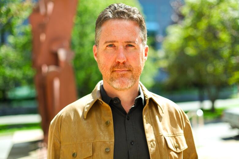 A man with brown-grey hair and a beard, wearing a tan blazer, stands outside.