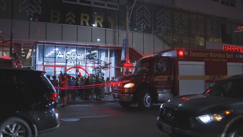 At night, on a city street, a fire truck is parked next to a sidewalk where a crowd of people are cordoned off by police tape