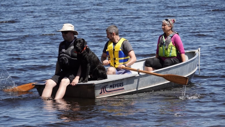 Three people sit in a boat with a black dog sitting in the front.