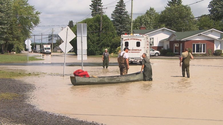 A person in a canoe floats in a dirty river on a street. People stand knee-deep in water. 