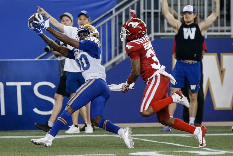 A football player wearing a blue and gold uniform catches a football while a player on the opposing team runs after them. 