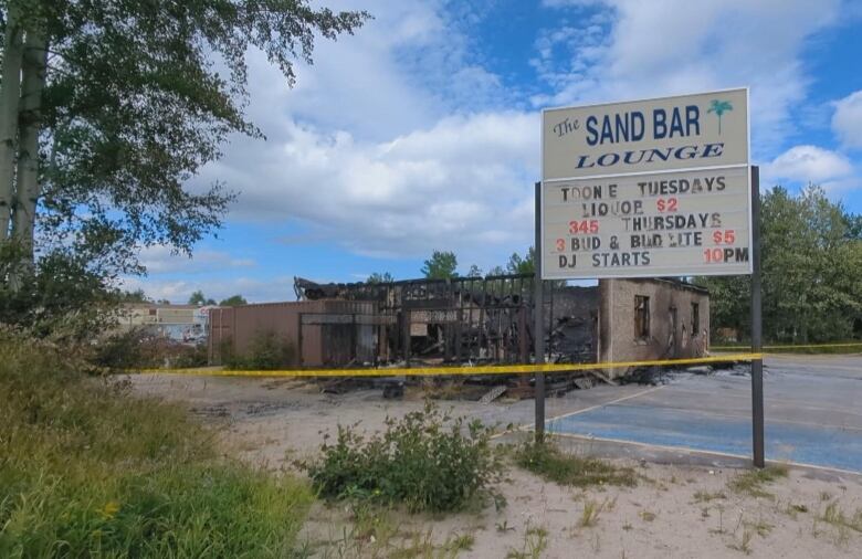 A sign saying Sand Bar lounge next to a burned out building with police tape accross it.