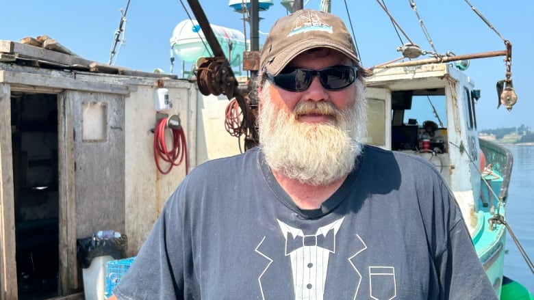 A white man with long white beard. Wearing a hart and shades. Standing in front of a fishing boat.