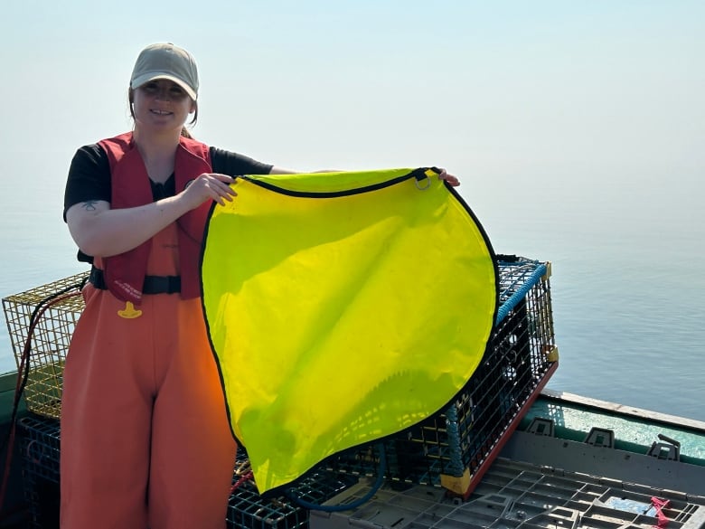 A white woman wearing a life jacket and a hat, holding up an inflatable balloon that mounts on an ropeless on-demand fishing system equipped with an air tank. She is standing on a fishing boat in water.