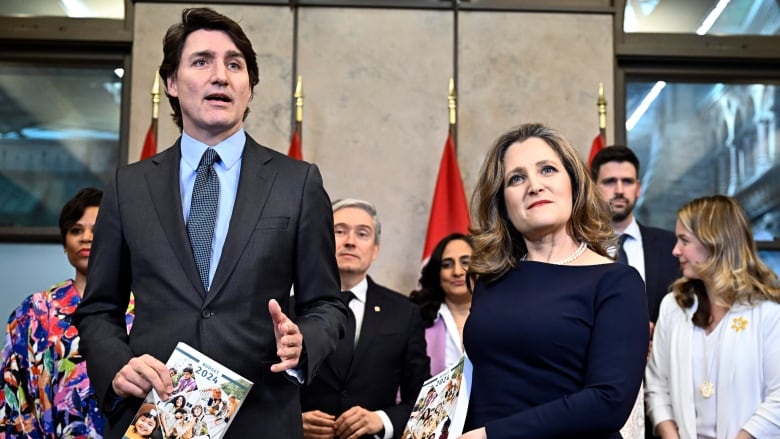 A man and a woman in formal attire stand together holding a catalogue like book. A group of others in business attire stand in the background.