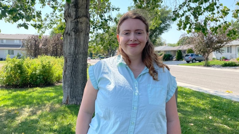 A woman in a blue button up shirt stands in a park. A tree is behind her. 