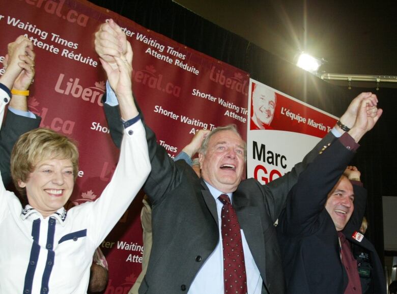 A man in a suit raises the arms of two people at his sides as they smile for an audience.