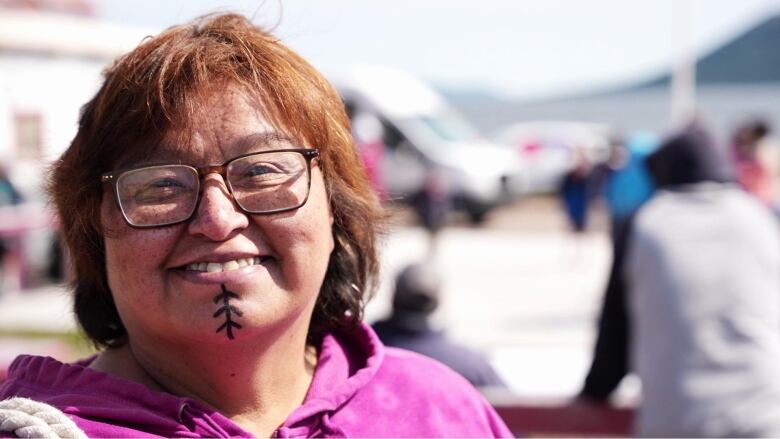 A woman in a pink sweater has a marking with a line and three arrows on her chin. 