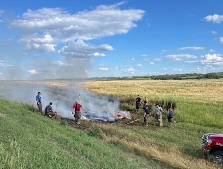 People with tools stand around a burning bale in a field during the day.