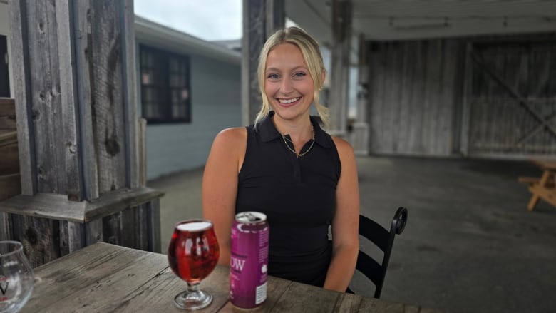 a woman smiles with a can of cider and a full glass of red cider in front of her