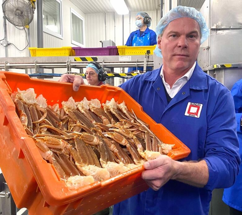 Man in hairnet and blue coat holds orange container full of crab legs.