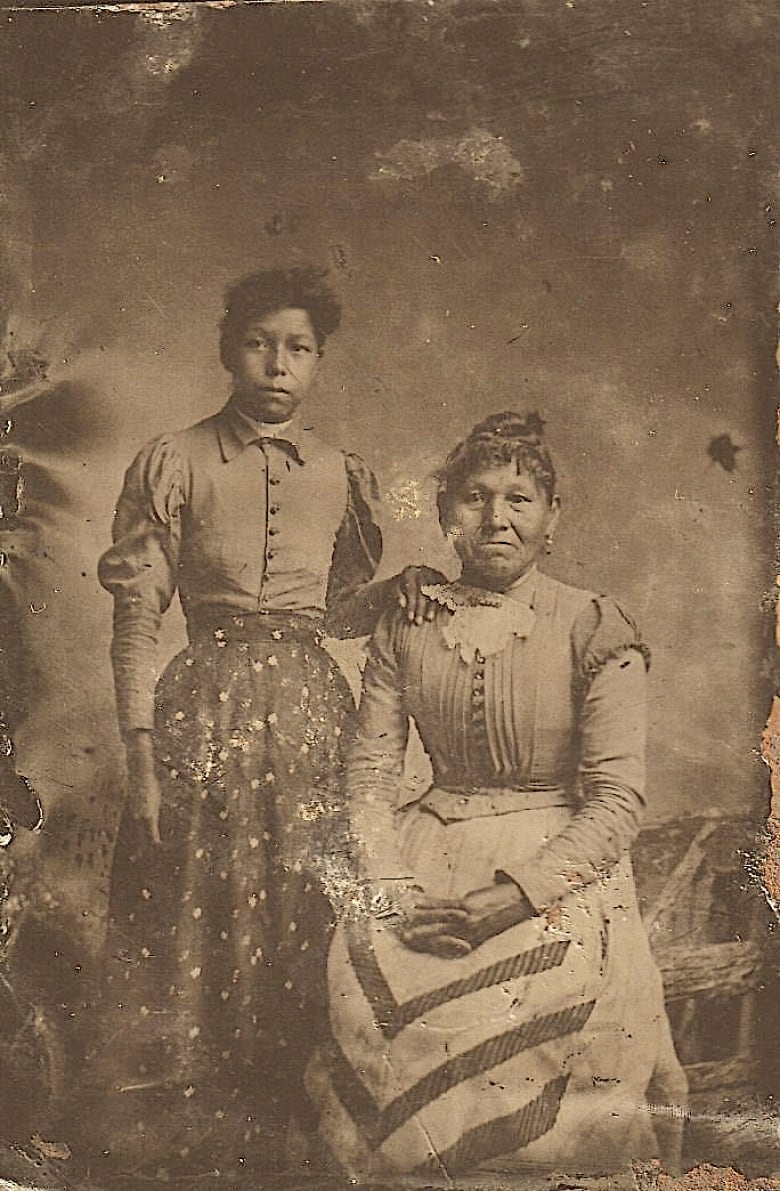 An old photographer shows a younger woman standing beside an older woman who is sitting.