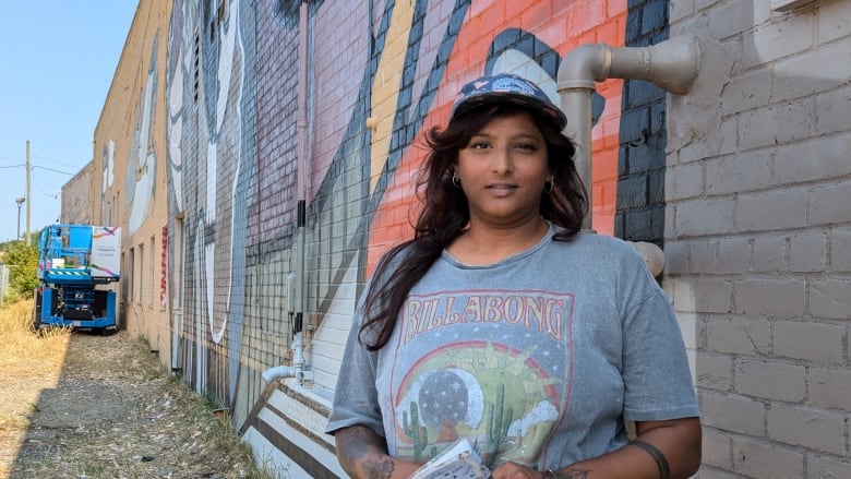 A woman wearing a graphic tee and a baseball cap standing in front of a mural.