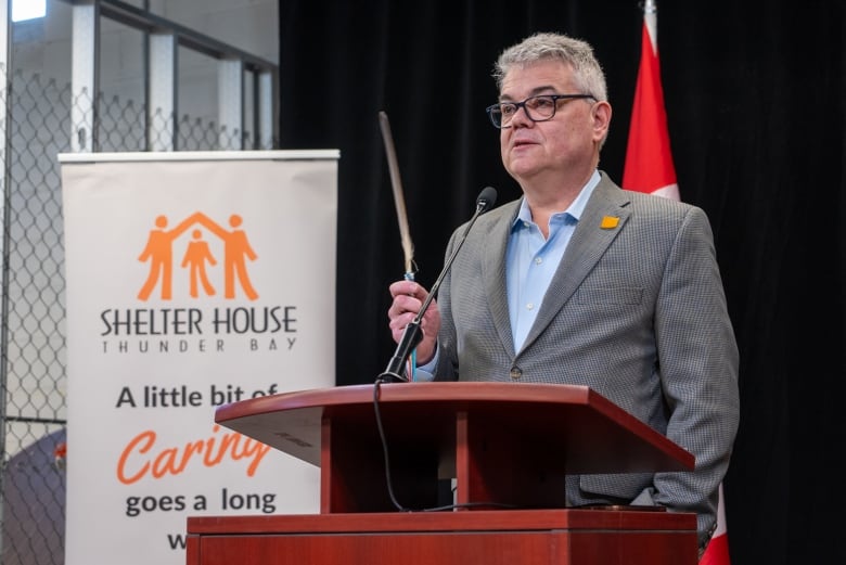 A person is seen standing at a podium and speaking into a microphone. They are holding a brown feather.