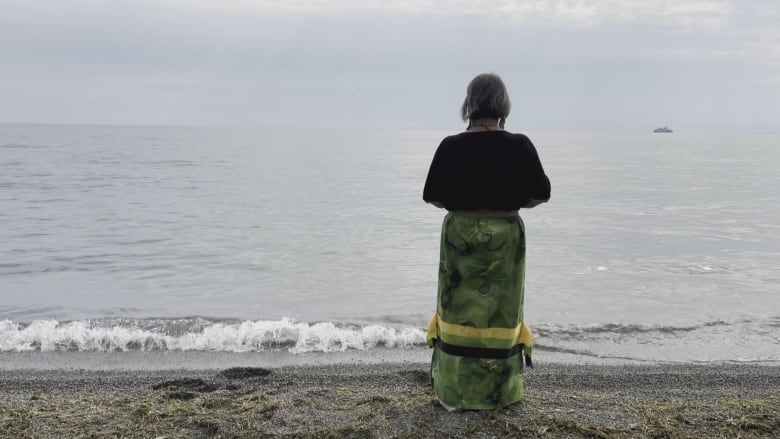 A woman stands facing a big body of water. Her back is to the camera.