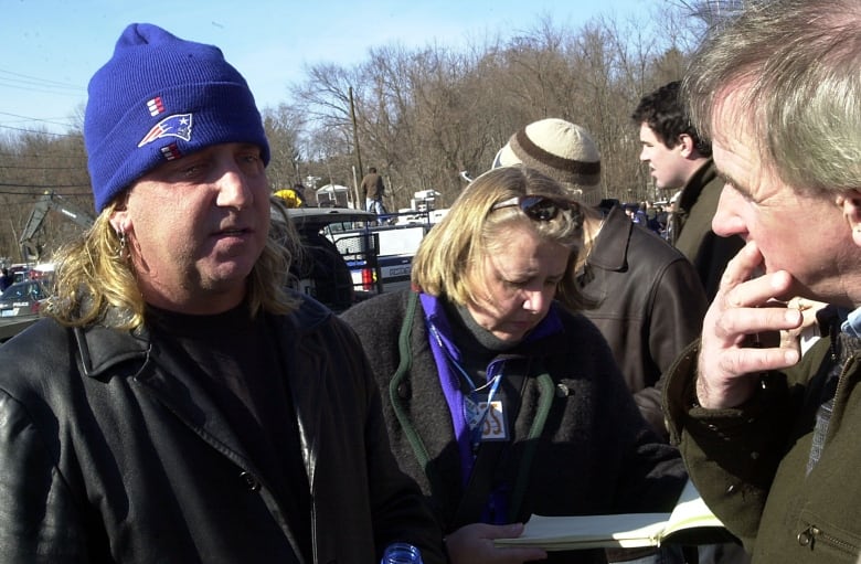 A man with long hair wearing a toque speaks outdoors while reporters gather around him.