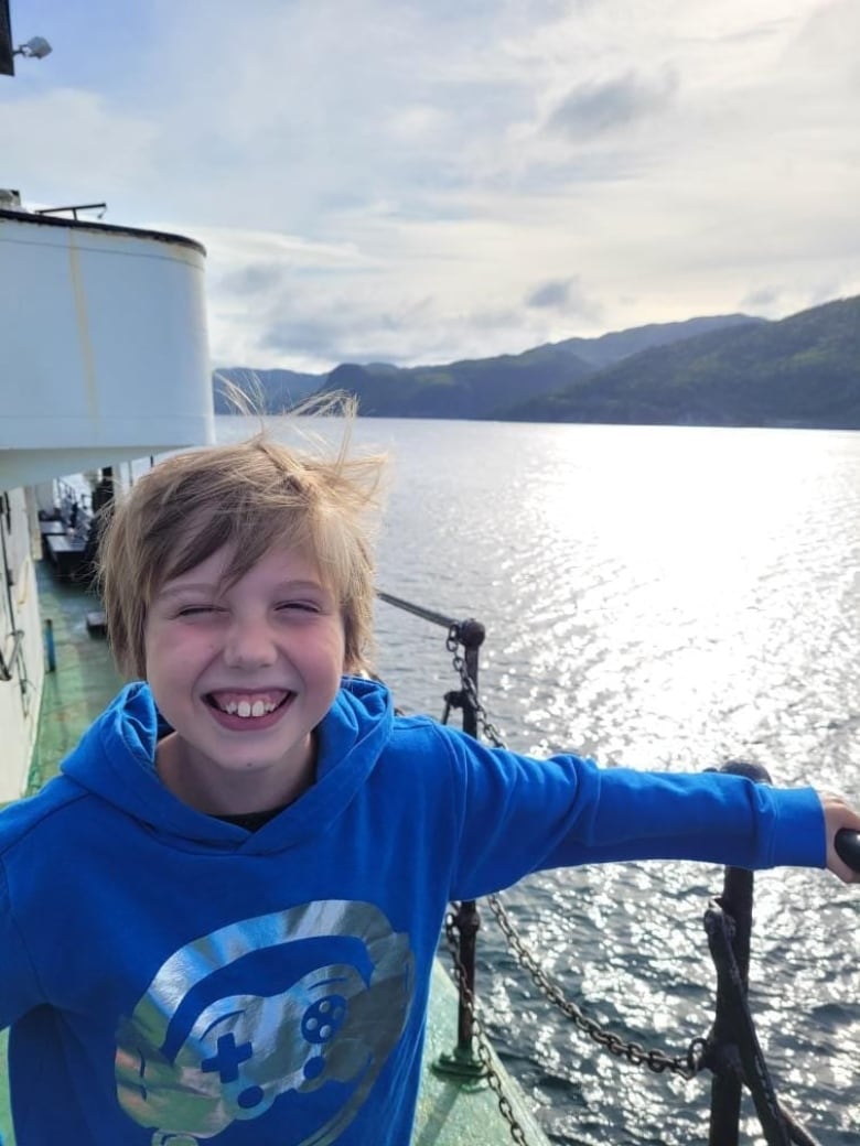 Young boy stands on boat smiling with squinty eyes. The ocean is in the background. 