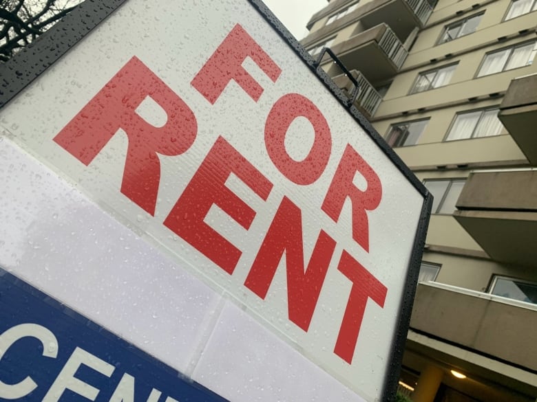 A sign with red letters reads FOR RENT. It's wet with rain, on an angle, and there's a beige highrise apartment building filling out the background.