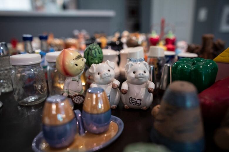 Dozens of salt and pepper shakers lay on a table. In focus are two melon-headed tennis player shaped ones, and two cute white pigs that are upright.